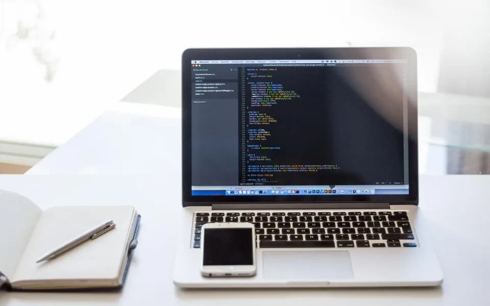 A laptop with code on screen on a bright desk beside a notebook.