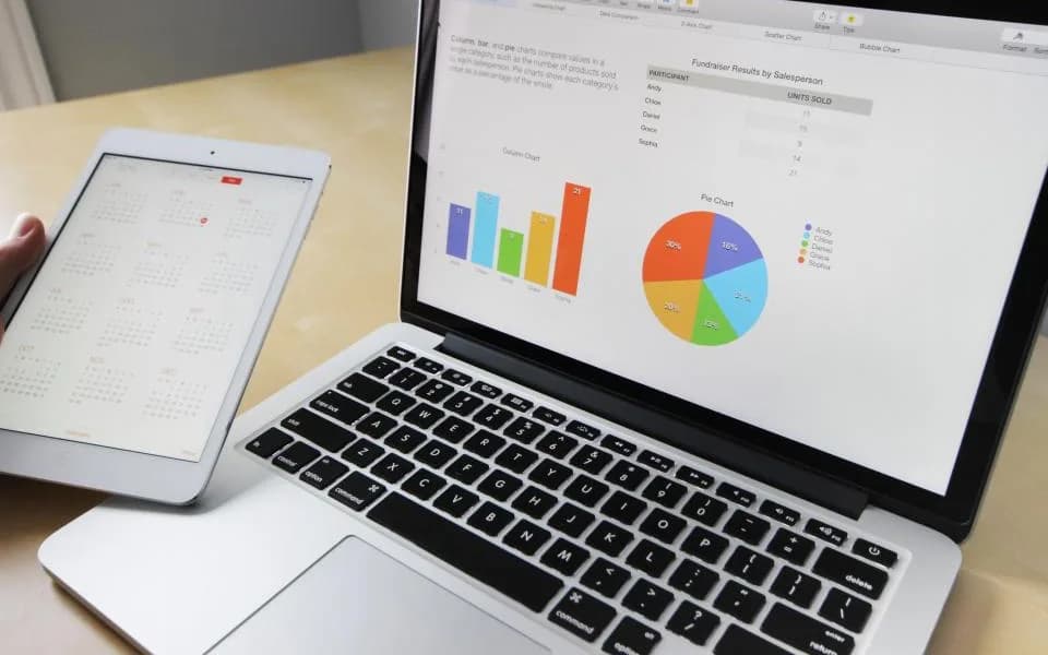 Bar and pie charts on a laptop beside a tablet on a desk.